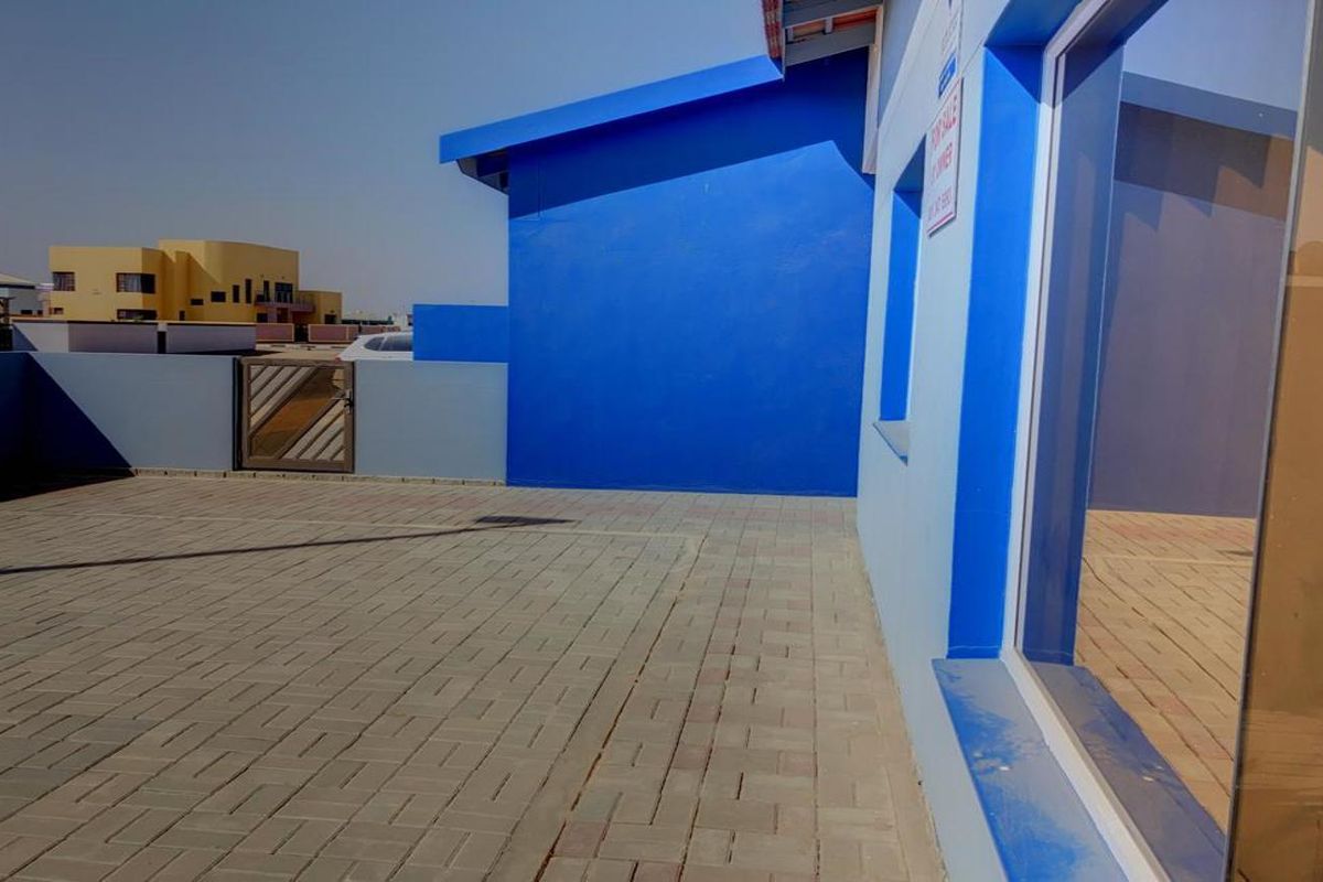 A paved rear patio area featuring a bright blue exterior wall, a metal gate, and a large sliding glass door providing access to the home.