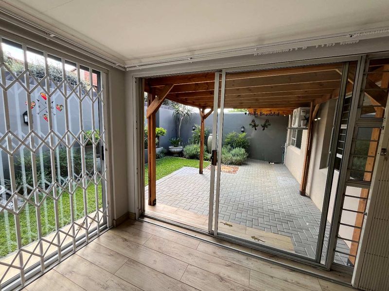 A bright, covered patio area featuring a wooden pergola, paved flooring, and direct access to a private garden space through sliding glass doors.