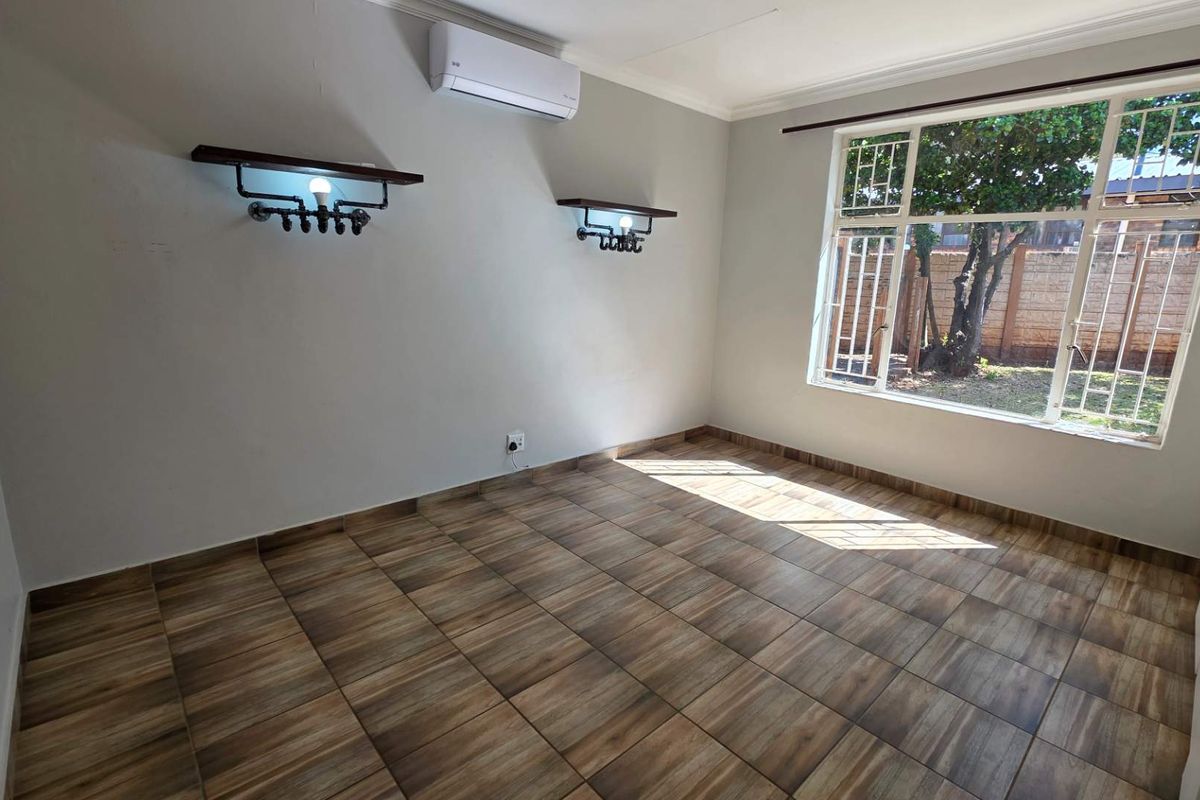 A bright, empty bedroom featuring wood-look tiled flooring, built-in cupboards, an air conditioning unit, and large windows with security bars.
