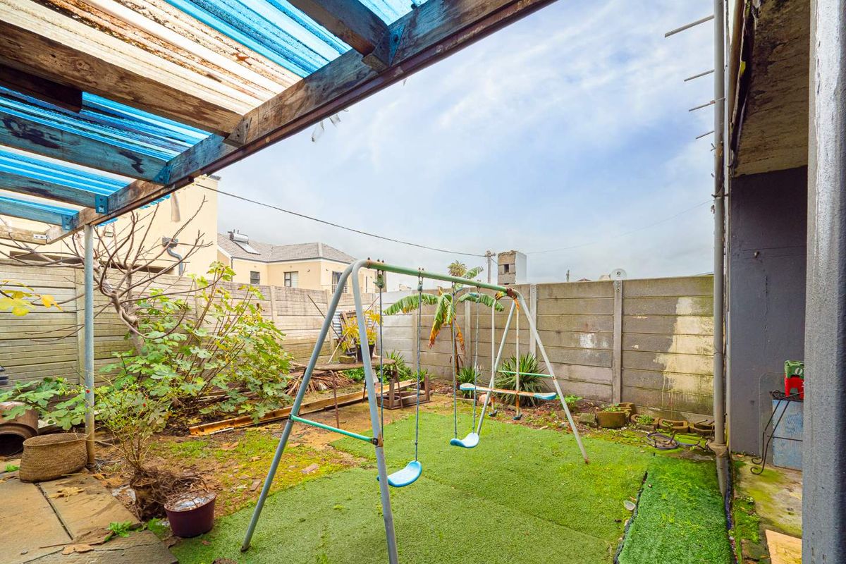 A rear garden area featuring a covered patio with a blue corrugated pergola, artificial lawn, and precast concrete boundary walls.