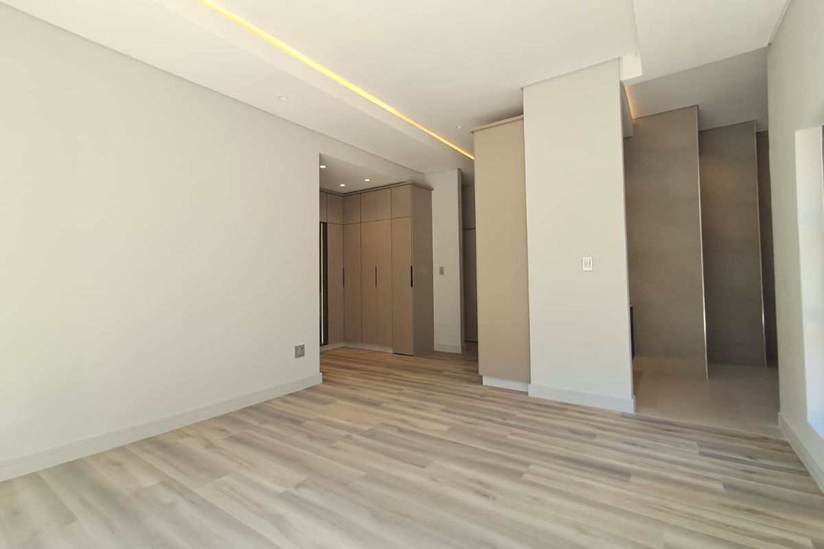 A modern, empty bedroom featuring light laminate flooring, neutral walls, built-in cupboards, and recessed ceiling lighting with ambient cove illumination.