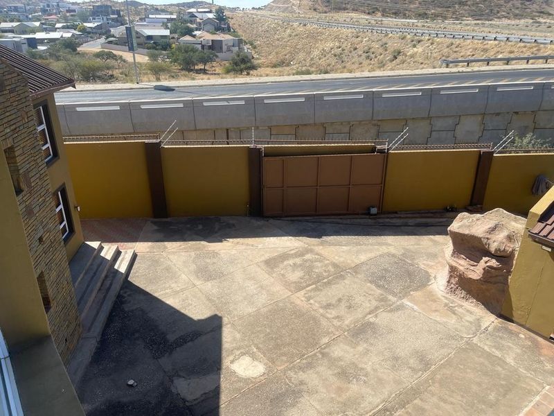 An elevated view of a paved rear courtyard featuring a sliding gate, perimeter wall with security wire, and stone-clad building exterior.