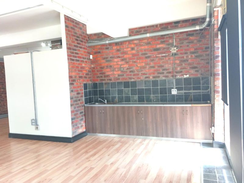 A compact office kitchenette featuring exposed brick walls, laminate flooring, built-in cabinetry, and a stainless steel sink with a tiled backsplash.
