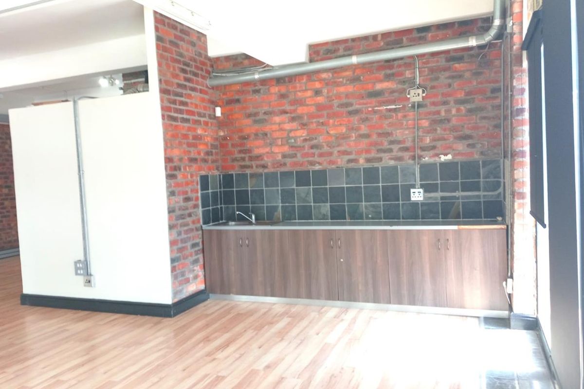 A compact office kitchenette featuring exposed brick walls, laminate flooring, built-in cabinetry, and a stainless steel sink with a tiled backsplash.