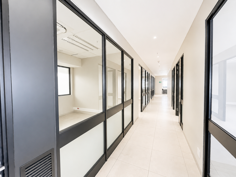 A modern, well-lit office hallway featuring clean tiled floors, glass partition walls, and a professional, minimalist aesthetic.