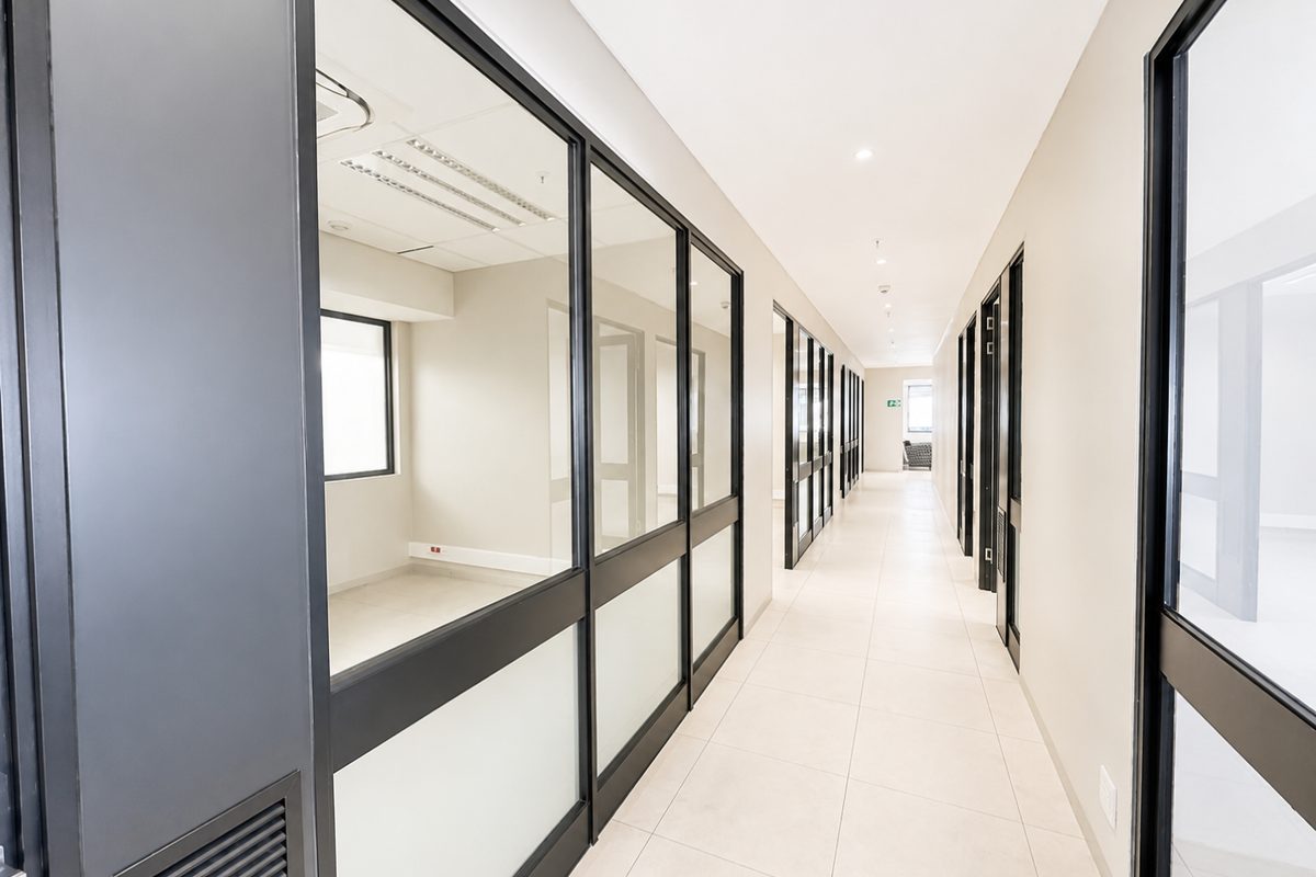 A modern, well-lit office hallway featuring clean tiled floors, glass partition walls, and a professional, minimalist aesthetic.
