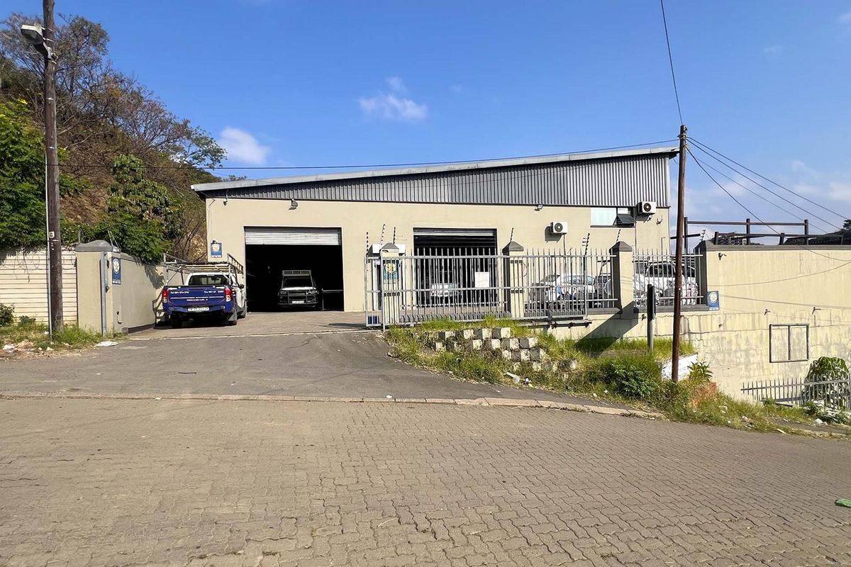 Industrial property featuring a warehouse with metal roller shutter doors, secure perimeter fencing, and an asphalt driveway entrance.