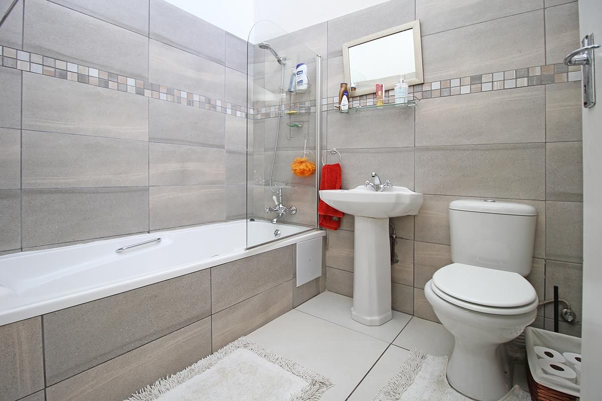 A modern bathroom featuring floor-to-ceiling grey tiling, a pedestal basin, a toilet, and a bathtub with a glass shower screen.