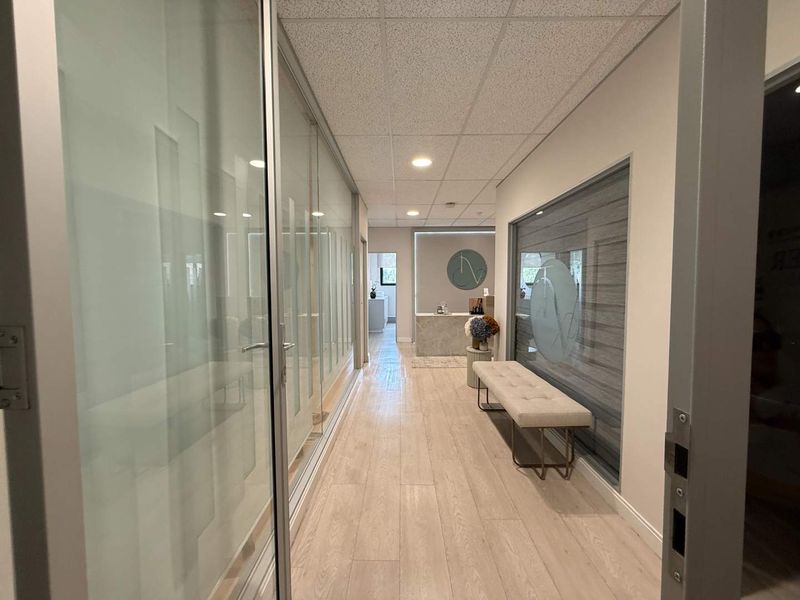 A modern office hallway featuring wood-look flooring, a suspended ceiling with recessed lighting, and a glass partition wall leading to a reception area.