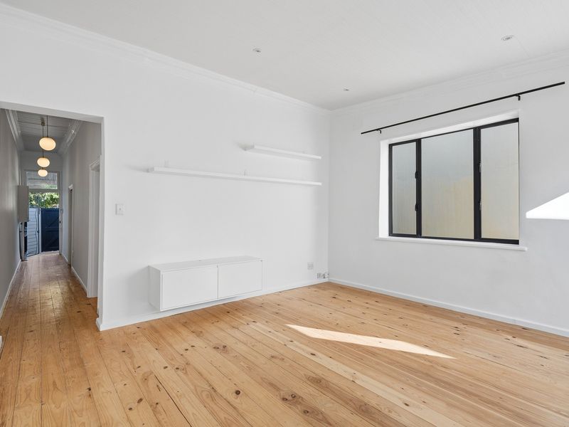 A bright, empty living room featuring polished timber flooring, built-in wall cabinetry, floating shelves, and a view through to the hallway.