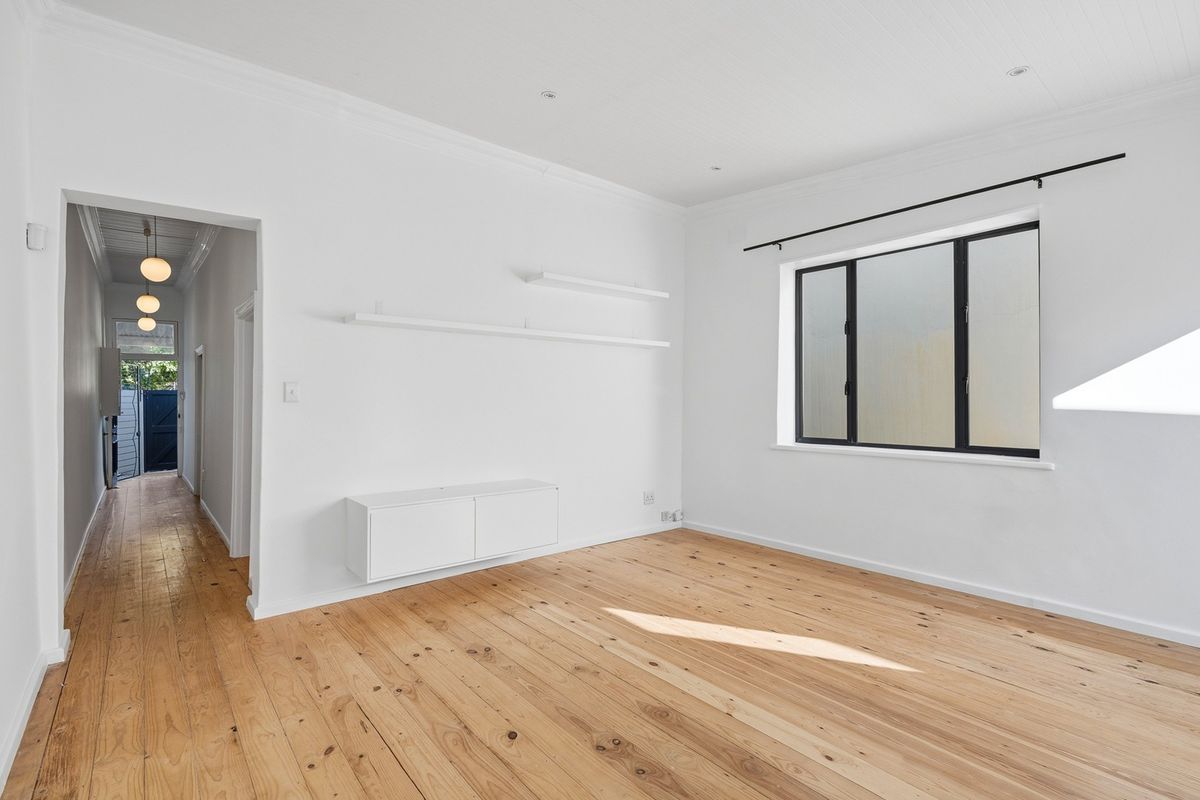 A bright, empty living room featuring polished timber flooring, built-in wall cabinetry, floating shelves, and a view through to the hallway.