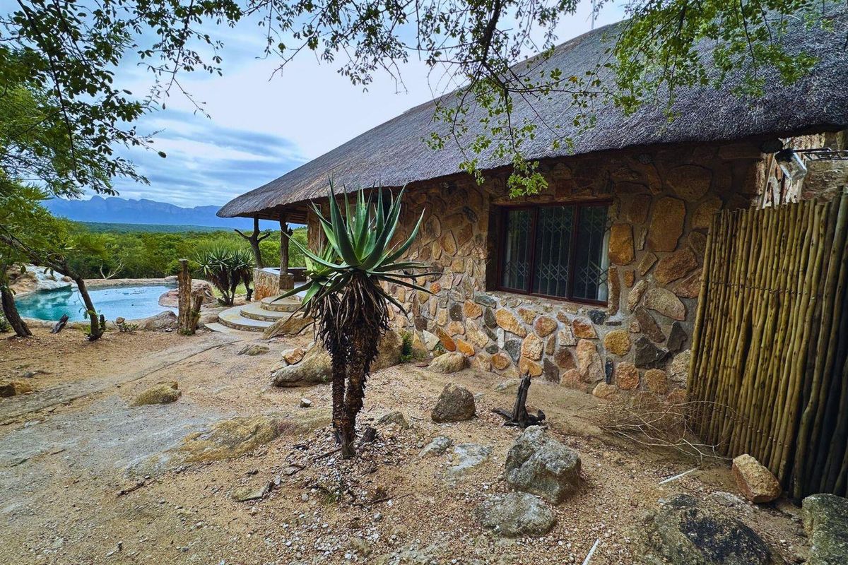 A rustic stone-clad house featuring a traditional thatched roof, set in a natural landscape with a swimming pool and mountain views.