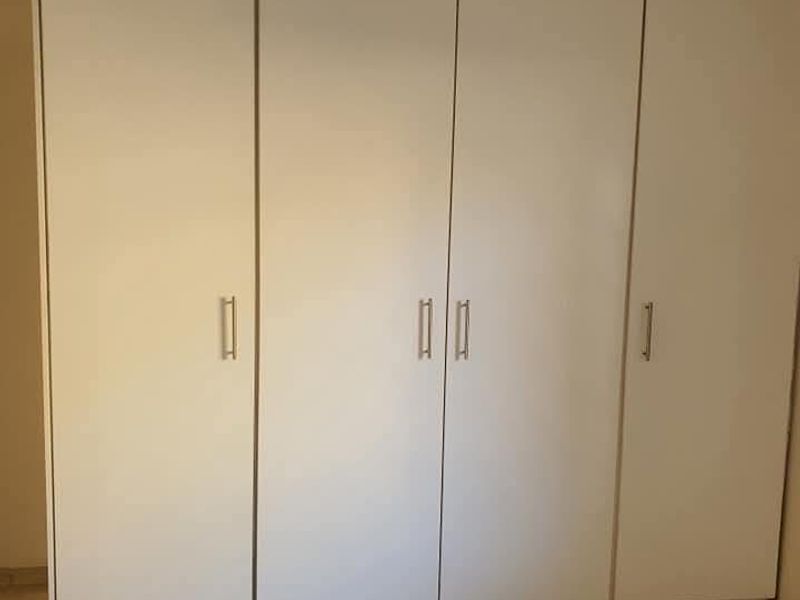 A clean bedroom interior featuring a large, white built-in wardrobe with modern handles and neutral tiled flooring.