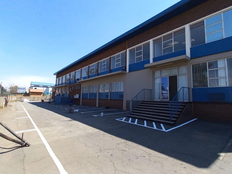 Front elevation of an industrial building featuring a paved parking area, brick facade, and multi-level office windows with metal frames.