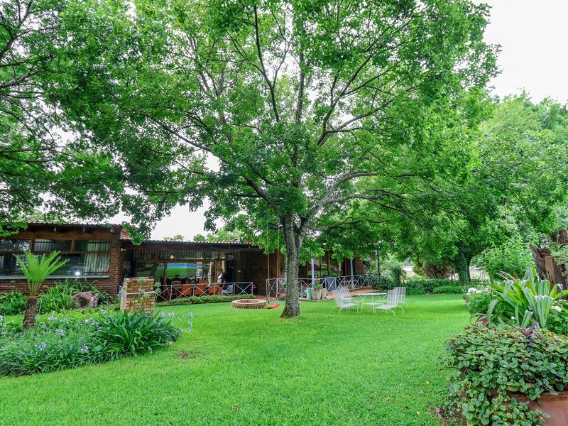 A spacious rear garden featuring a well-maintained lawn, mature trees, a brick fire pit, and a covered patio area attached to the brick farmhouse.
