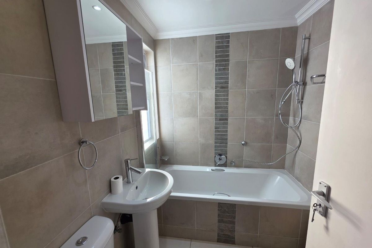 A modern, well-maintained bathroom featuring neutral wall tiles, a pedestal basin, a built-in bathtub with shower attachment, and a mirrored medicine cabinet.