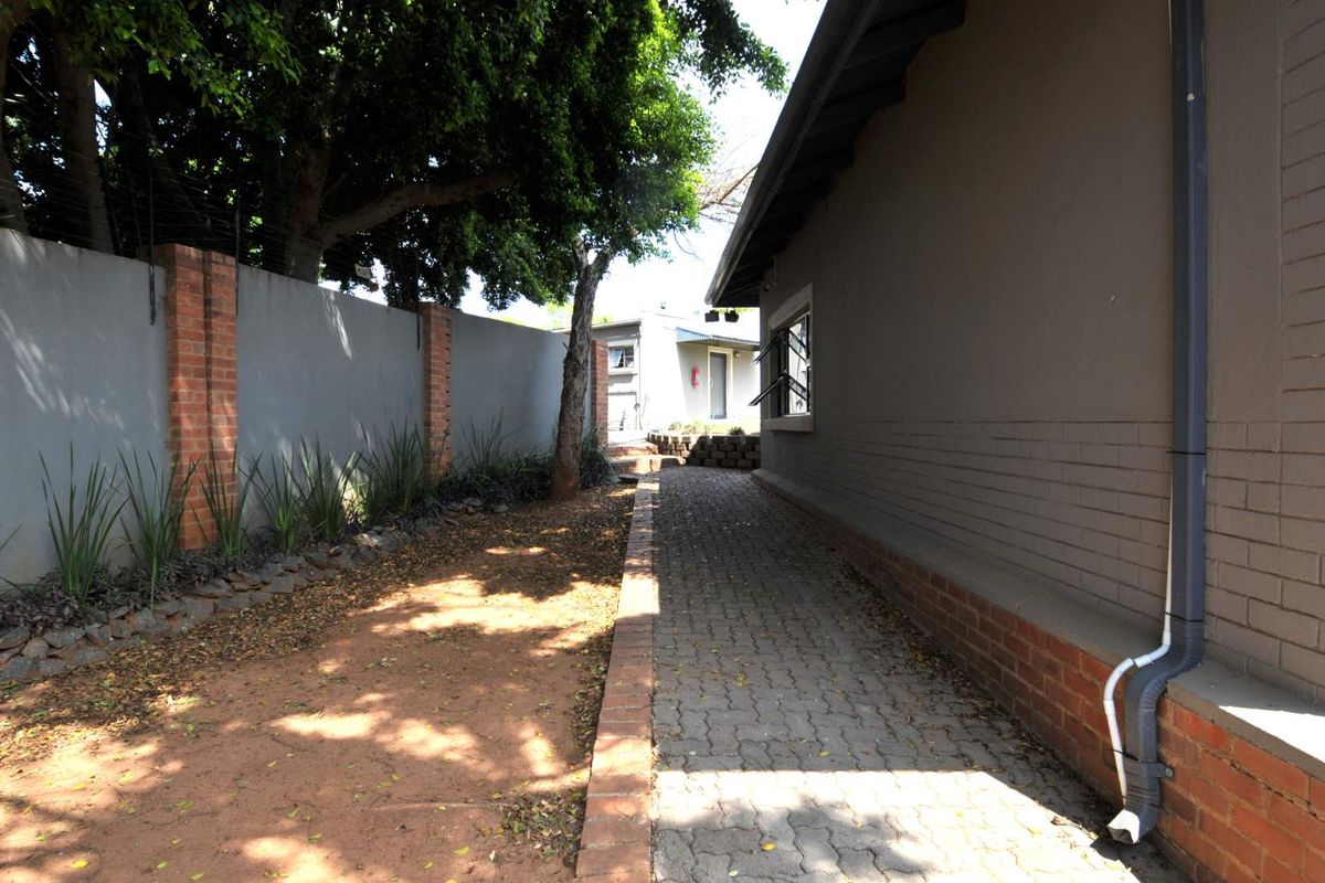 A side exterior view showing a brick paved walkway alongside the building, bordered by a perimeter wall and mature trees.