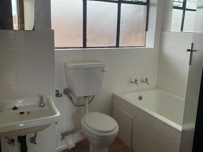A dated bathroom featuring a built-in bathtub, pedestal basin, and flush toilet, with tiled finishes and a steel-framed window.