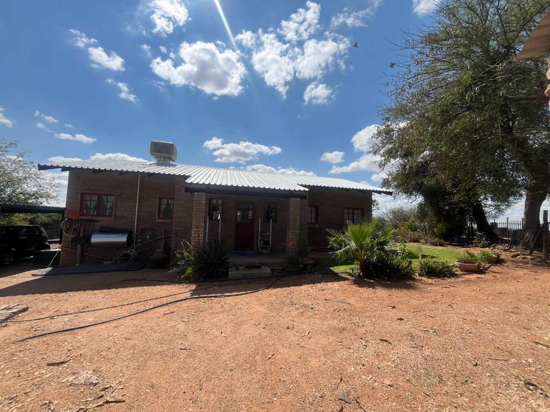 A single-story brick house featuring a corrugated metal roof, an evaporative cooling unit, and a covered front porch, set on a large unpaved plot.