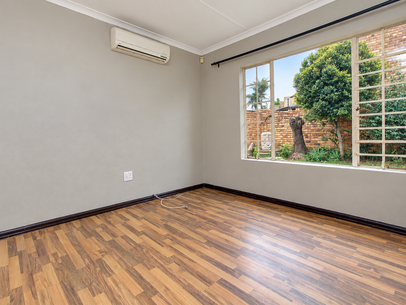 A bright, empty bedroom featuring laminate flooring, a wall-mounted air conditioning unit, and large windows with security bars overlooking the garden.