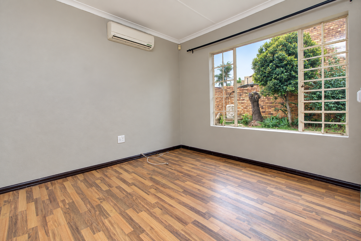 A bright, empty bedroom featuring laminate flooring, a wall-mounted air conditioning unit, and large windows with security bars overlooking the garden.