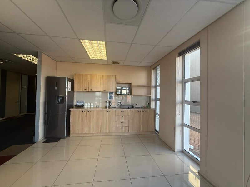A functional office kitchenette featuring light wood-finish cabinetry, tiled flooring, a stainless steel sink, and ample natural light from a large window.