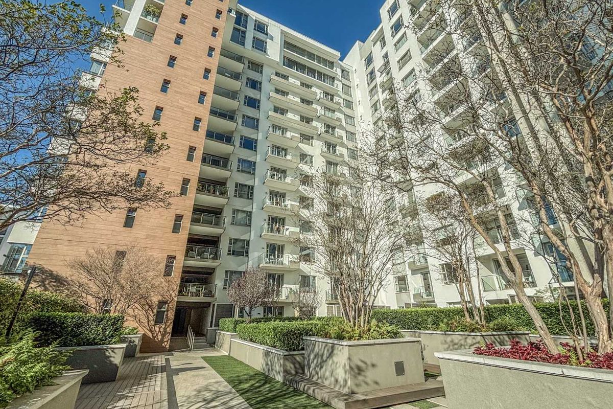 A view of the apartment building exterior featuring landscaped garden beds, paved walkways, and multiple levels of private balconies.