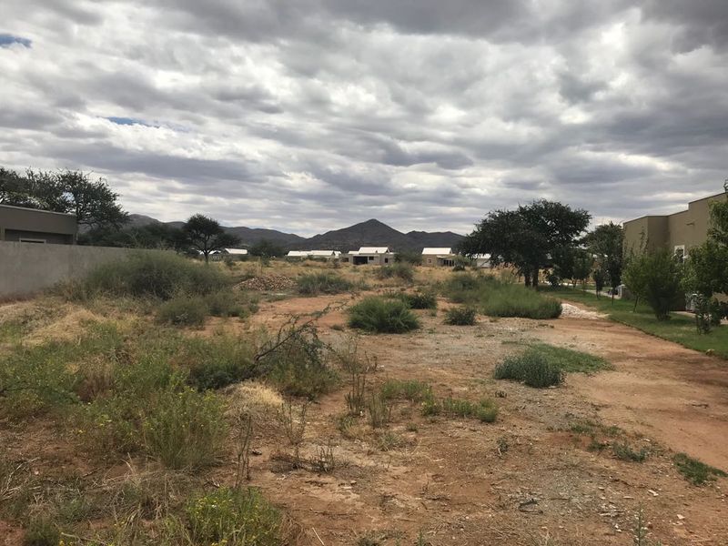 A view of a vacant residential plot featuring natural vegetation, a boundary wall, and surrounding residential developments in the distance.