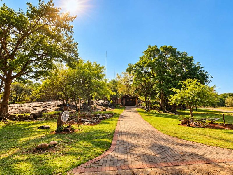 A scenic paved walkway leading through a well-maintained garden with mature trees and natural rock formations towards the property entrance.