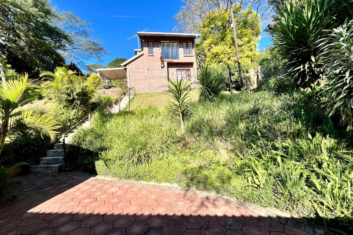 A rear view of a multi-story brick house featuring a paved patio area, concrete stairs with handrails, and a lush, sloped garden.