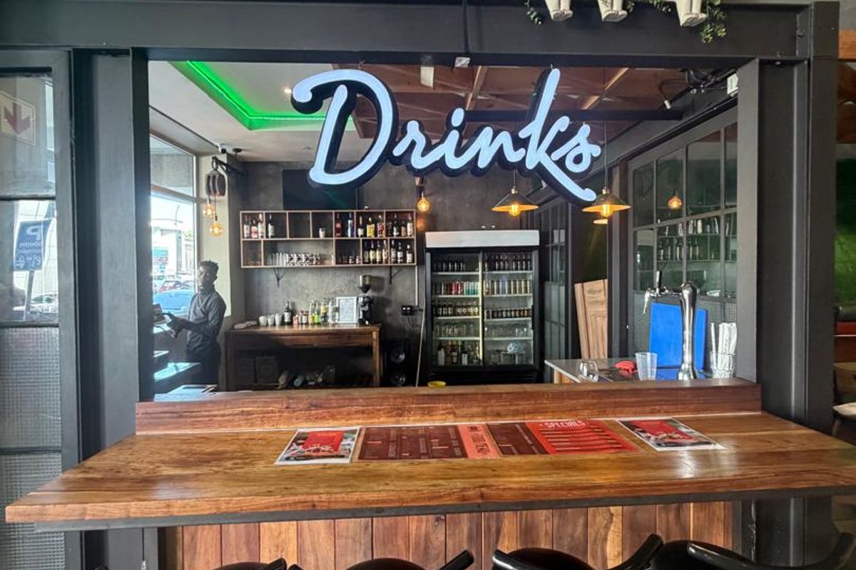 A well-appointed bar area featuring a wooden service counter, industrial-style steel framing, integrated shelving, and a dedicated drinks display fridge.