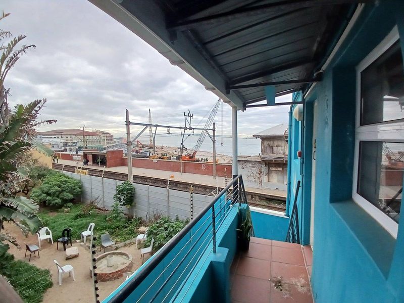 An elevated balcony featuring tiled flooring and a metal balustrade, overlooking a garden area with views of the nearby railway and coastline.