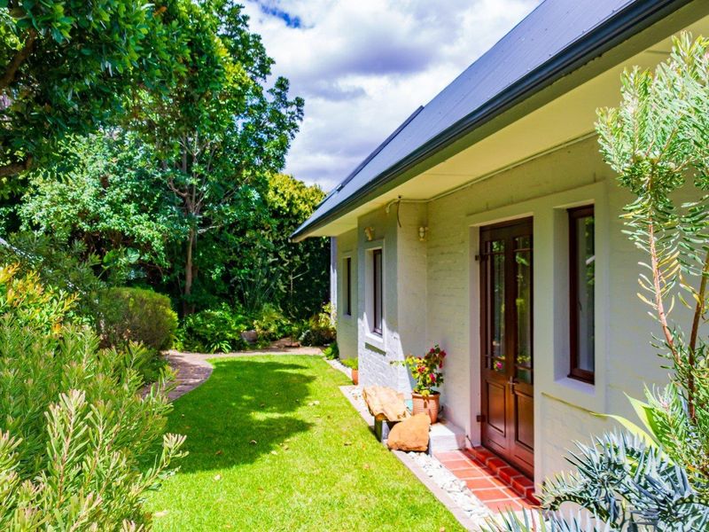 A well-maintained rear exterior view featuring a brick facade, metal roof, timber door, and a lush, landscaped garden with a paved walkway.
