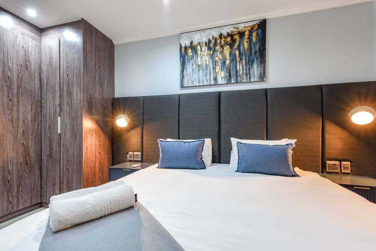 A modern, well-appointed bedroom featuring a large upholstered headboard, integrated wardrobe cabinetry, and stylish wall-mounted lighting fixtures.