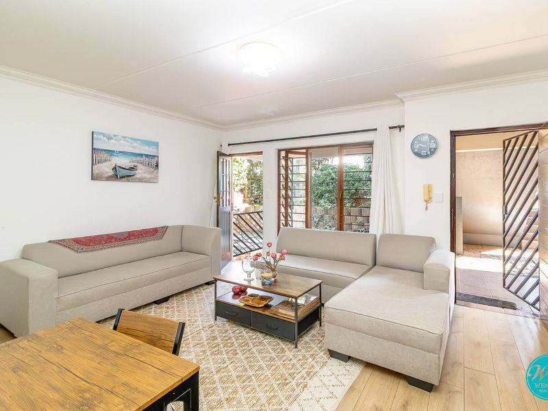 A bright living room featuring laminate flooring, timber-framed windows, and direct access to an outdoor area through a security gate.