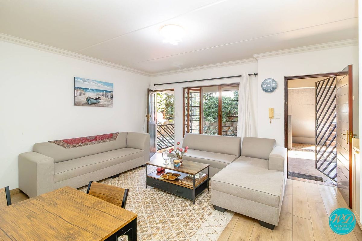 A bright living room featuring laminate flooring, timber-framed windows, and direct access to an outdoor area through a security gate.