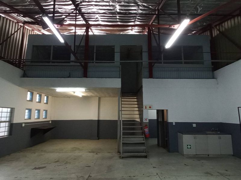 Industrial warehouse space featuring a mezzanine level, metal staircase, exposed roof trusses, and a small kitchenette area with built-in cabinetry.