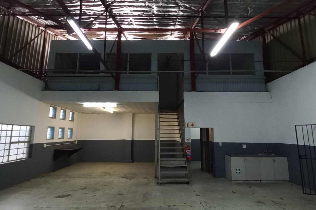 Industrial warehouse space featuring a mezzanine level, metal staircase, exposed roof trusses, and a small kitchenette area with built-in cabinetry.