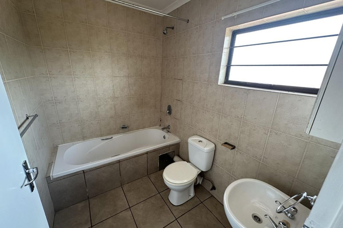 A functional bathroom featuring a built-in bathtub with shower head, pedestal basin, and toilet, finished with neutral wall and floor tiling.