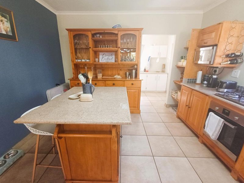 A functional kitchen featuring wooden cabinetry, a central island, built-in oven and hob, with an adjoining scullery area and tiled flooring throughout.