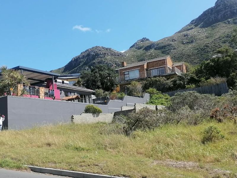 A view of a vacant residential plot with a sloped landscape, boundary walls, and surrounding established residential properties against a mountain backdrop.