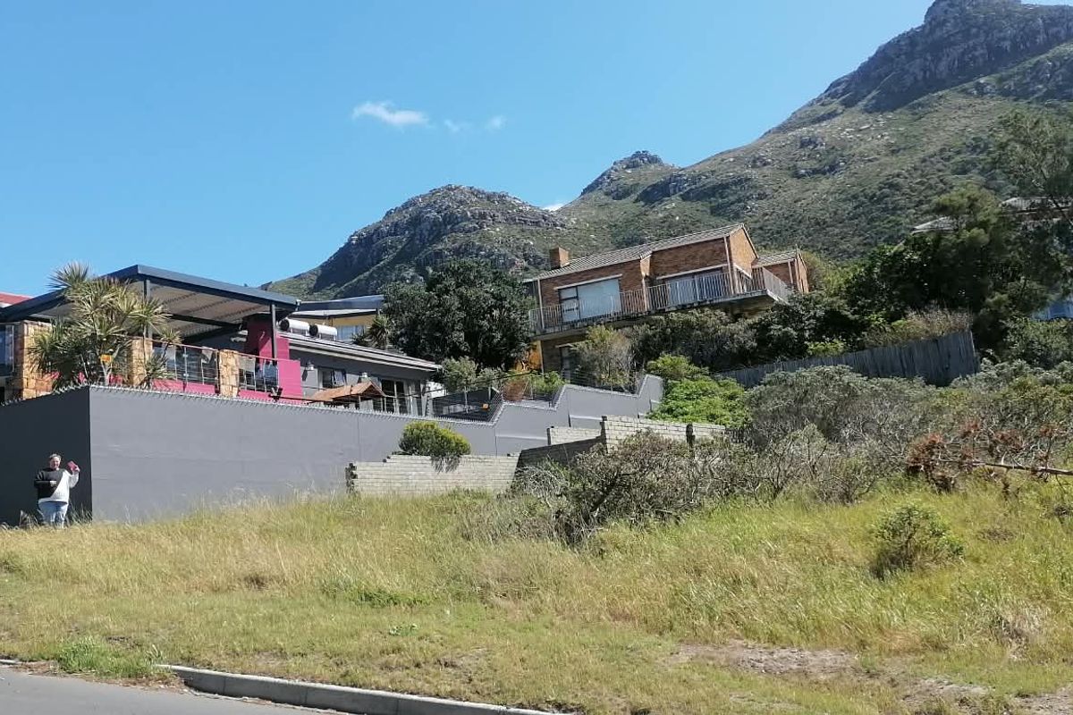 A view of a vacant residential plot with a sloped landscape, boundary walls, and surrounding established residential properties against a mountain backdrop.