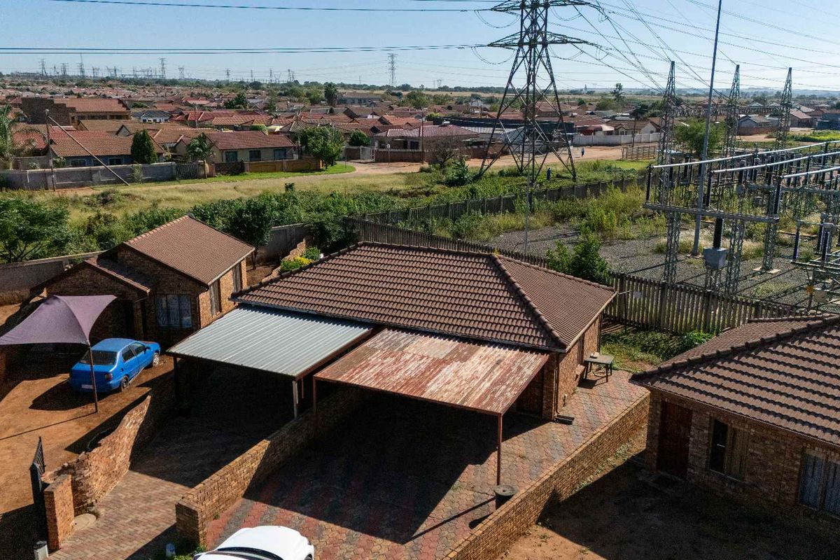 Aerial view of a townhouse property featuring a brick facade, tiled roof, and double carport structure with adjacent residential neighborhood and electrical infrastructure.