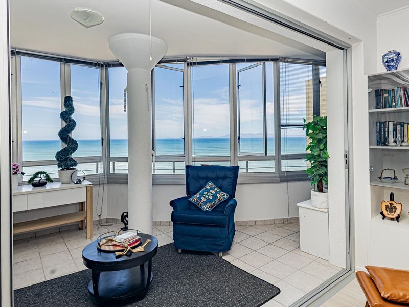 A bright, enclosed balcony featuring tiled flooring, a structural column, and expansive windows offering panoramic ocean views, seamlessly connected to the main living area.