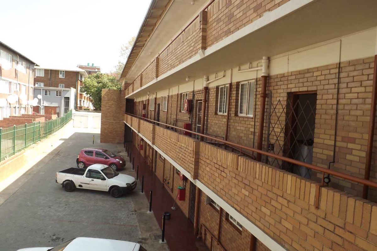 Exterior view of a multi-story apartment building featuring brick facade, external walkways, and metal security railings overlooking a paved parking area.