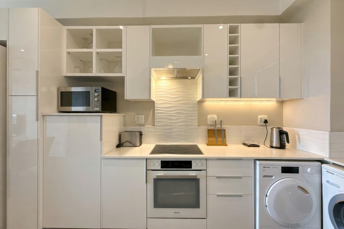 Modern kitchen featuring white high-gloss cabinetry, integrated oven, hob, extractor fan, and built-in laundry appliances with a textured tiled splashback.