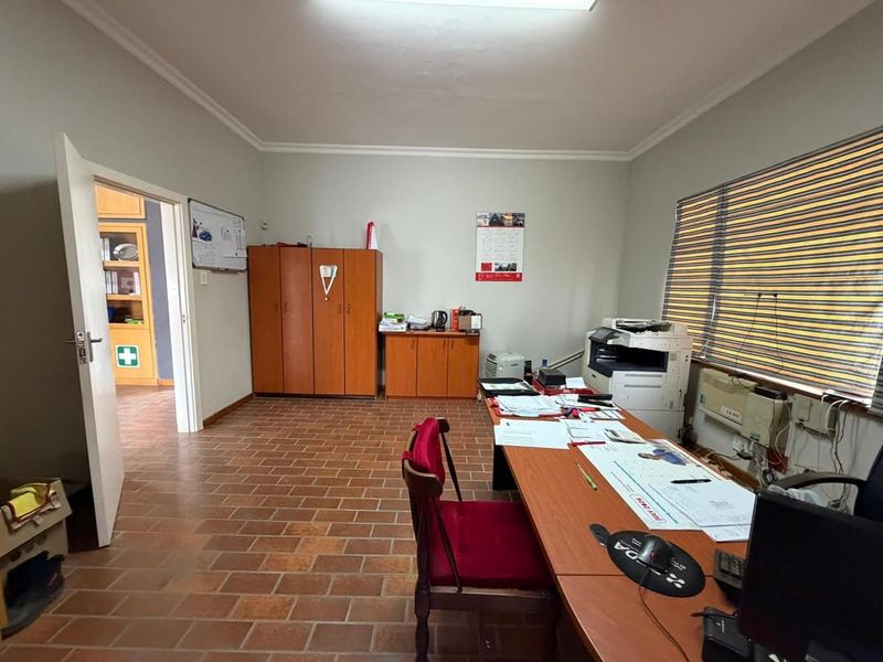 A functional office space featuring terracotta tile flooring, built-in storage cabinetry, and large windows with fitted blinds.