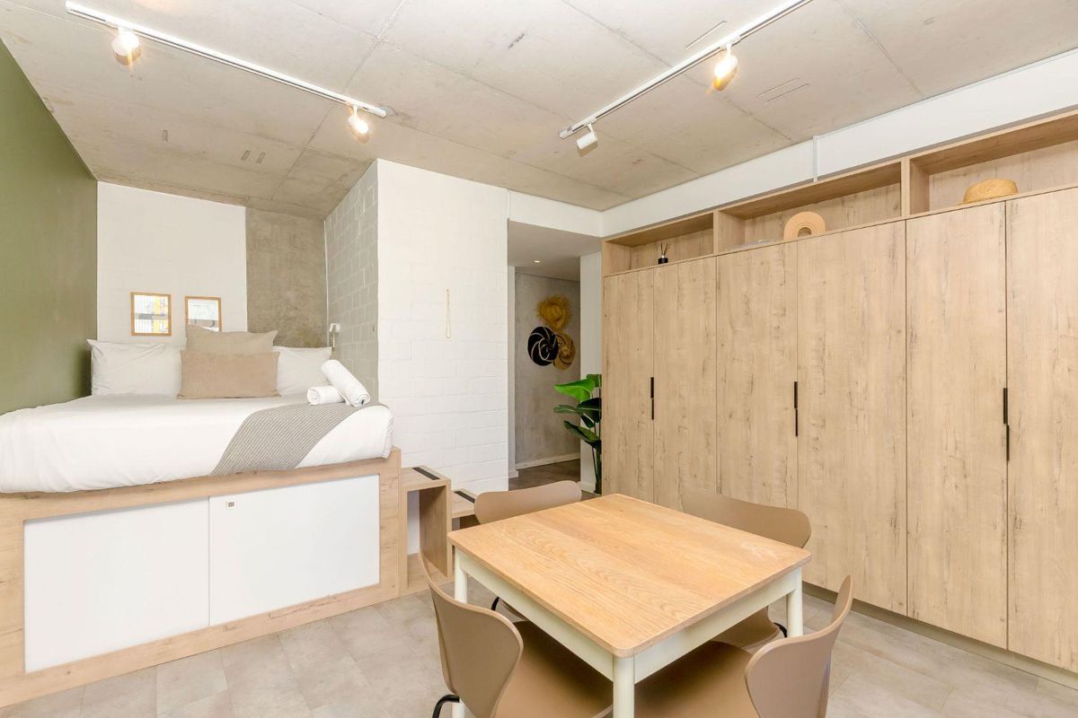 Modern bedroom featuring an exposed concrete ceiling, track lighting, a raised bed platform, and extensive built-in timber-look cabinetry for storage.