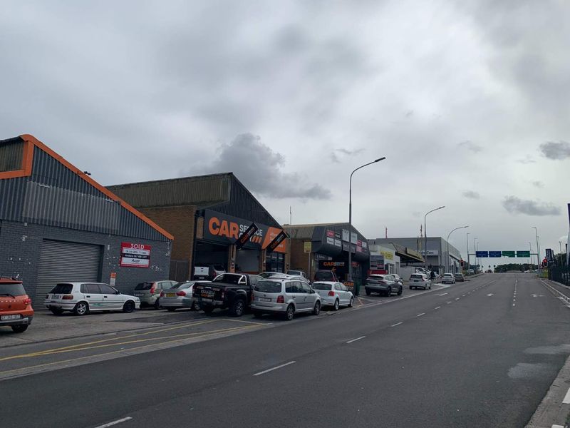 Street-level view of a commercial retail and industrial strip featuring multiple units with roller shutter doors and signage along a public road.