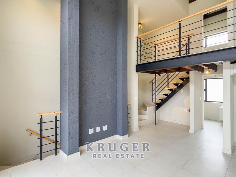Spacious open-plan living area featuring a double-volume ceiling, a striking dark feature wall, and a modern mezzanine level with a steel staircase.